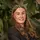 Zaza Jenkin, Designer at Plant Plan, smiling with long, wavy brown hair styled with gold hair clips. She is wearing a dark blouse and stands against a lush green plant backdrop, creating a natural and professional setting.