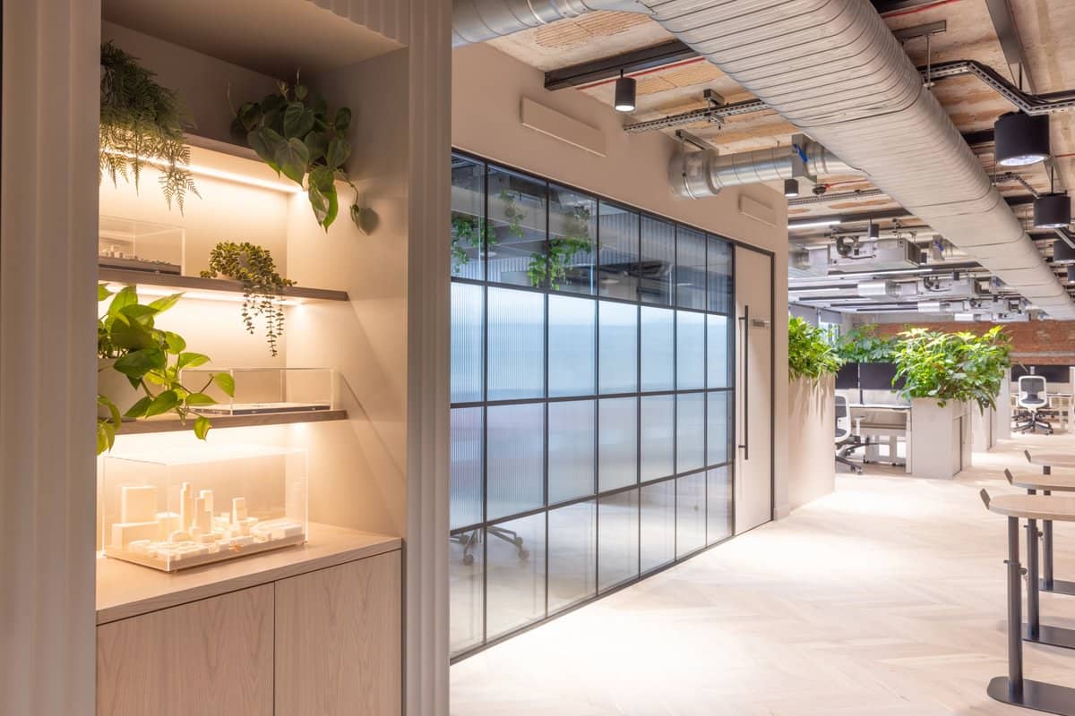 Modern office interior with glass partitions, wooden flooring, overhead ducts, and plants. Shelving with small architectural models on the left.