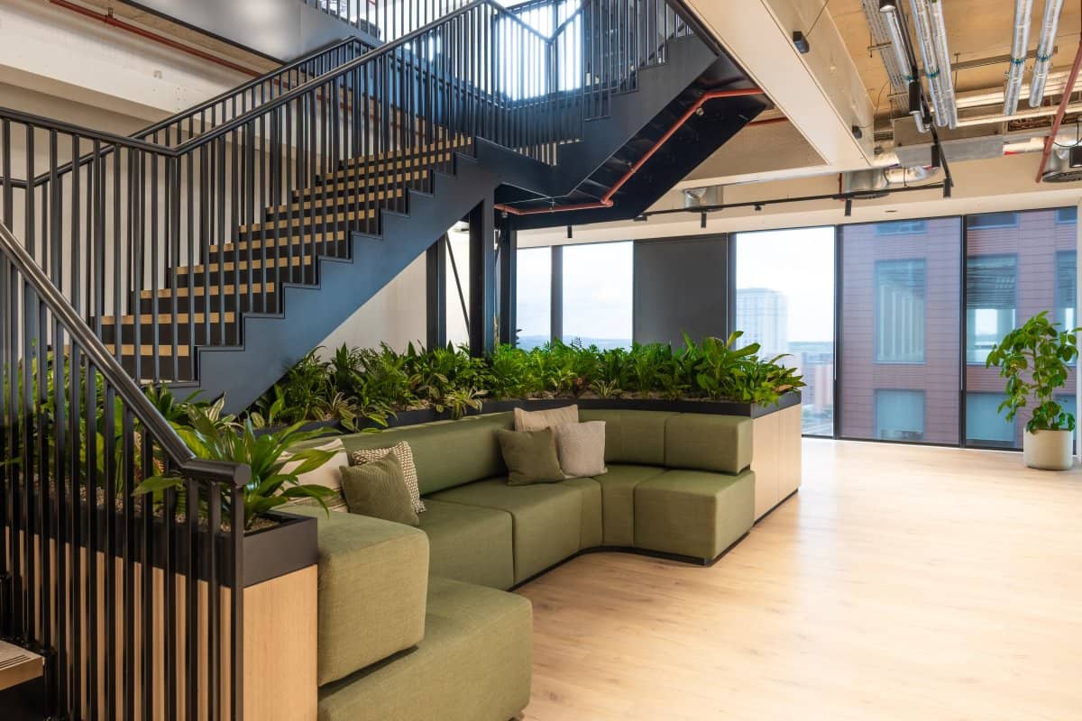 Modern office lobby with a green sectional sofa, indoor plants, and a metal staircase. Large windows let in natural light.