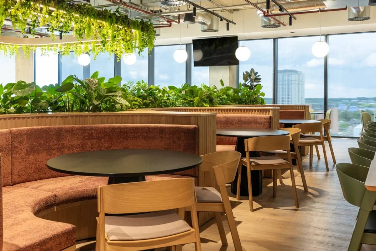Workspace breakout area with large windows, round tables, wooden chairs, hanging greenery, and plants. Overhead lights illuminate the cozy space.