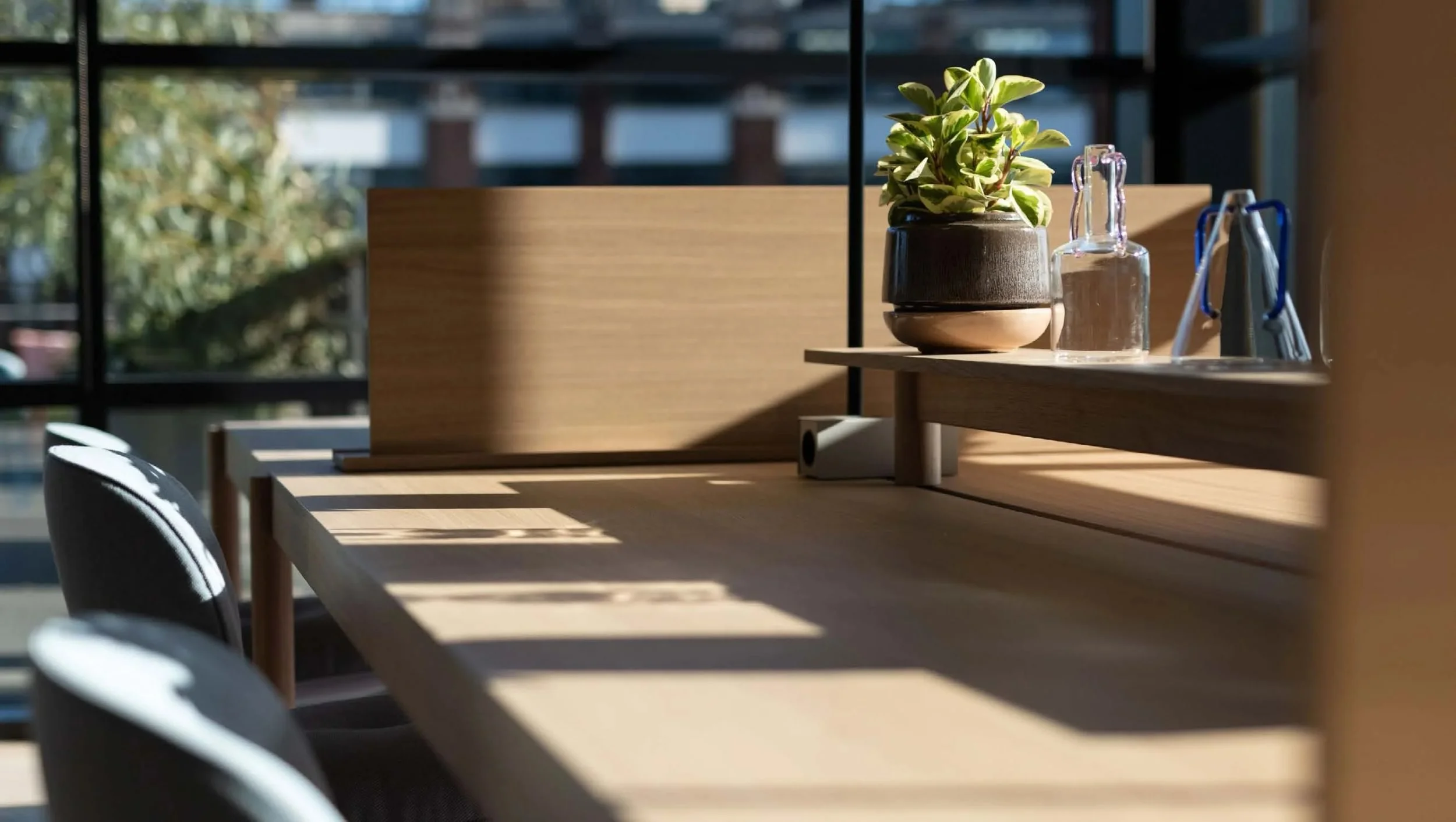 Sunlit desk with a small potted plant, a clear glass bottle, and napkins, casting shadows in a modern office setting.