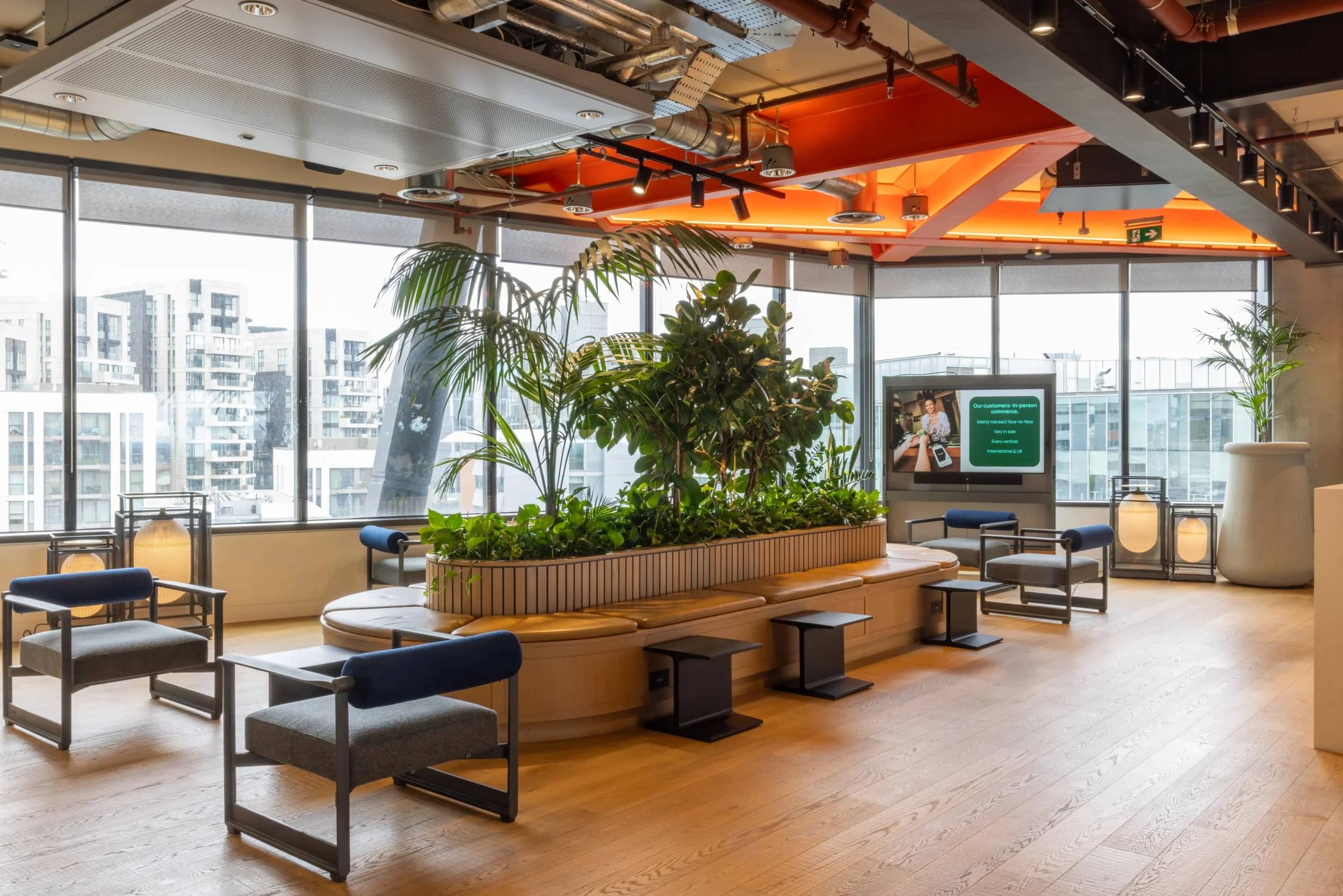 Modern office lounge with large windows, greenery, wooden floor, stylish chairs, and a digital screen. Bright, airy, and inviting space.