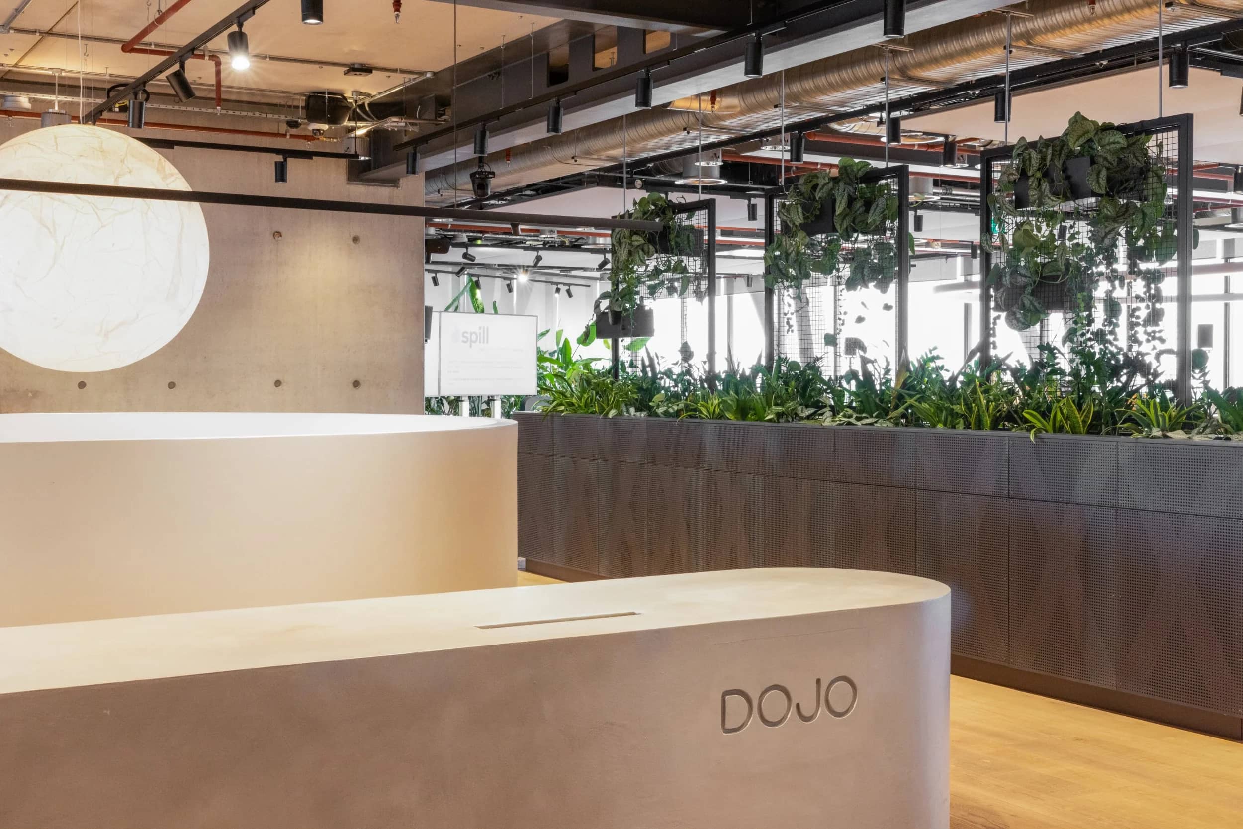 Modern office lobby with a minimalist desk labeled "DOJO," a large round light, and lush greenery in the background.