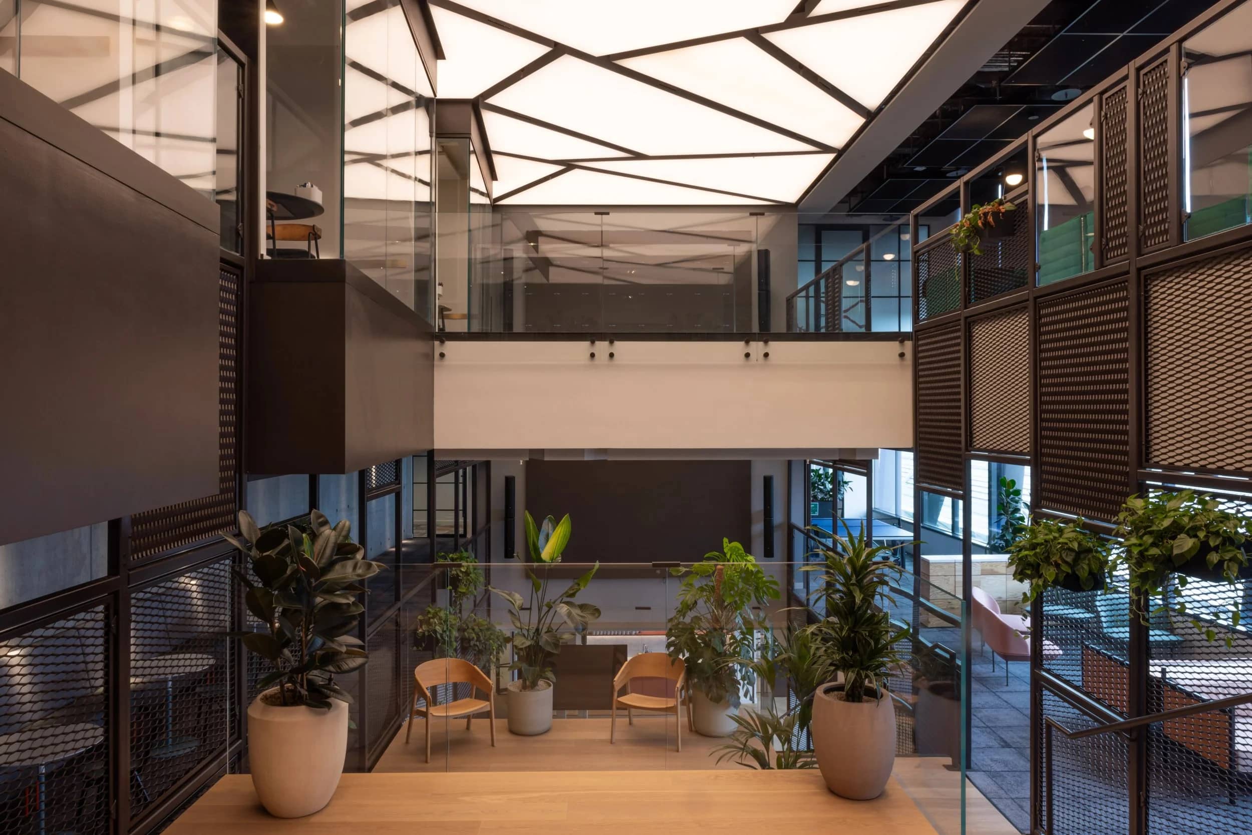 Modern office lobby with geometric ceiling lights, potted plants, glass railings, wooden floor, and seating area, creating a sleek, airy atmosphere.