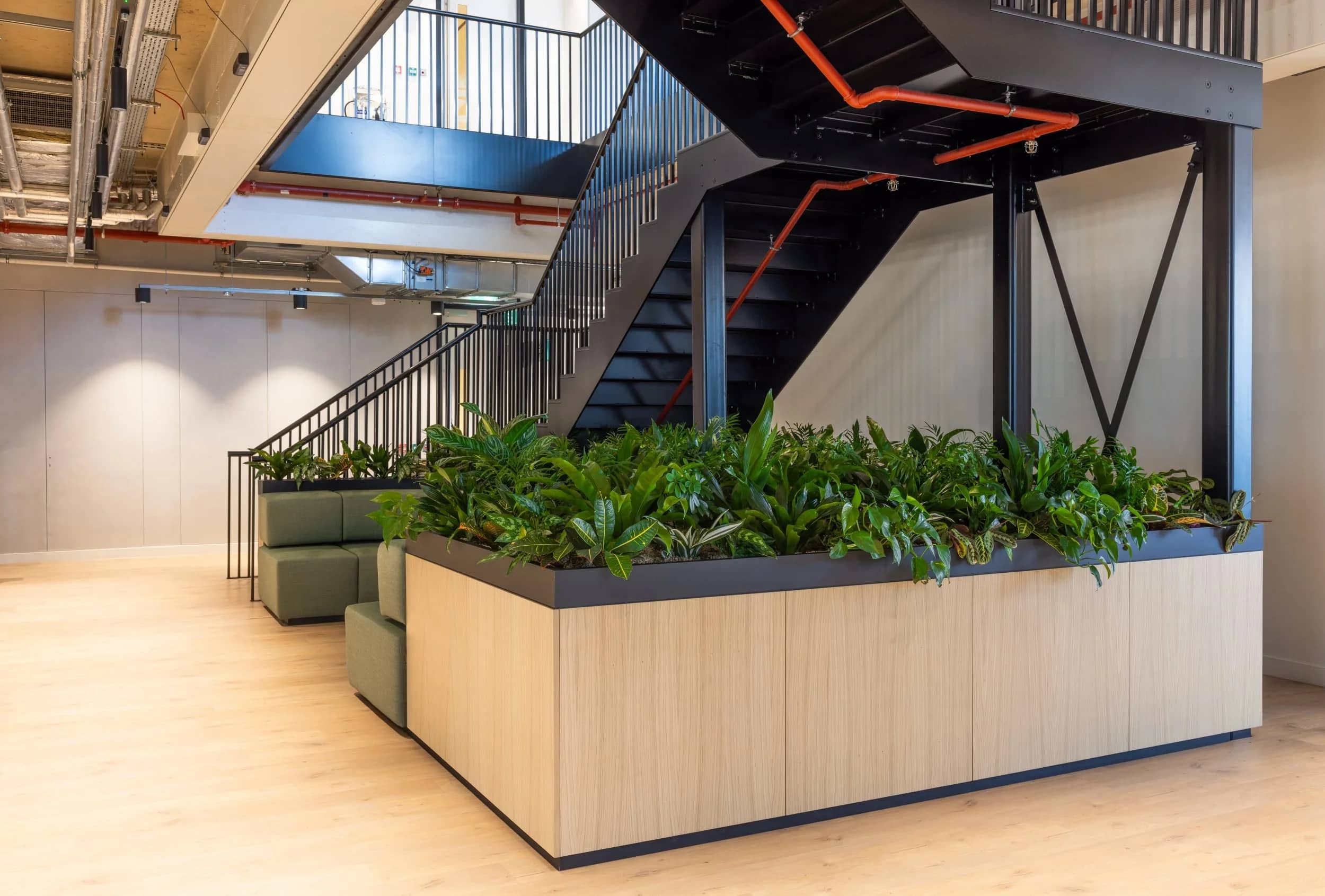 Modern office interior with a black metal staircase, wooden planters filled with green plants, and green seating on light wood flooring.