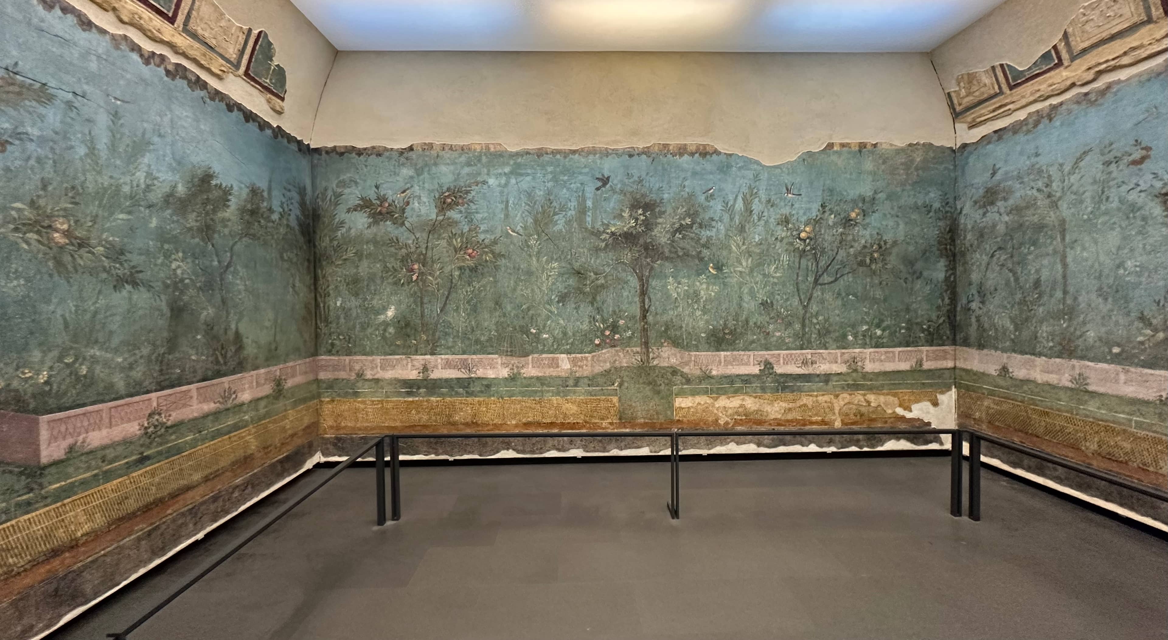 Ancient Roman fresco depicting a lush garden with trees, flowers, and a blue sky, displayed in a three-walled room.