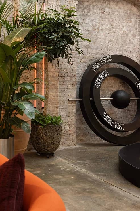 A modern interior with exposed brick walls, lush potted plants, and a large black circular sculpture featuring words like "Design" and "Culture."