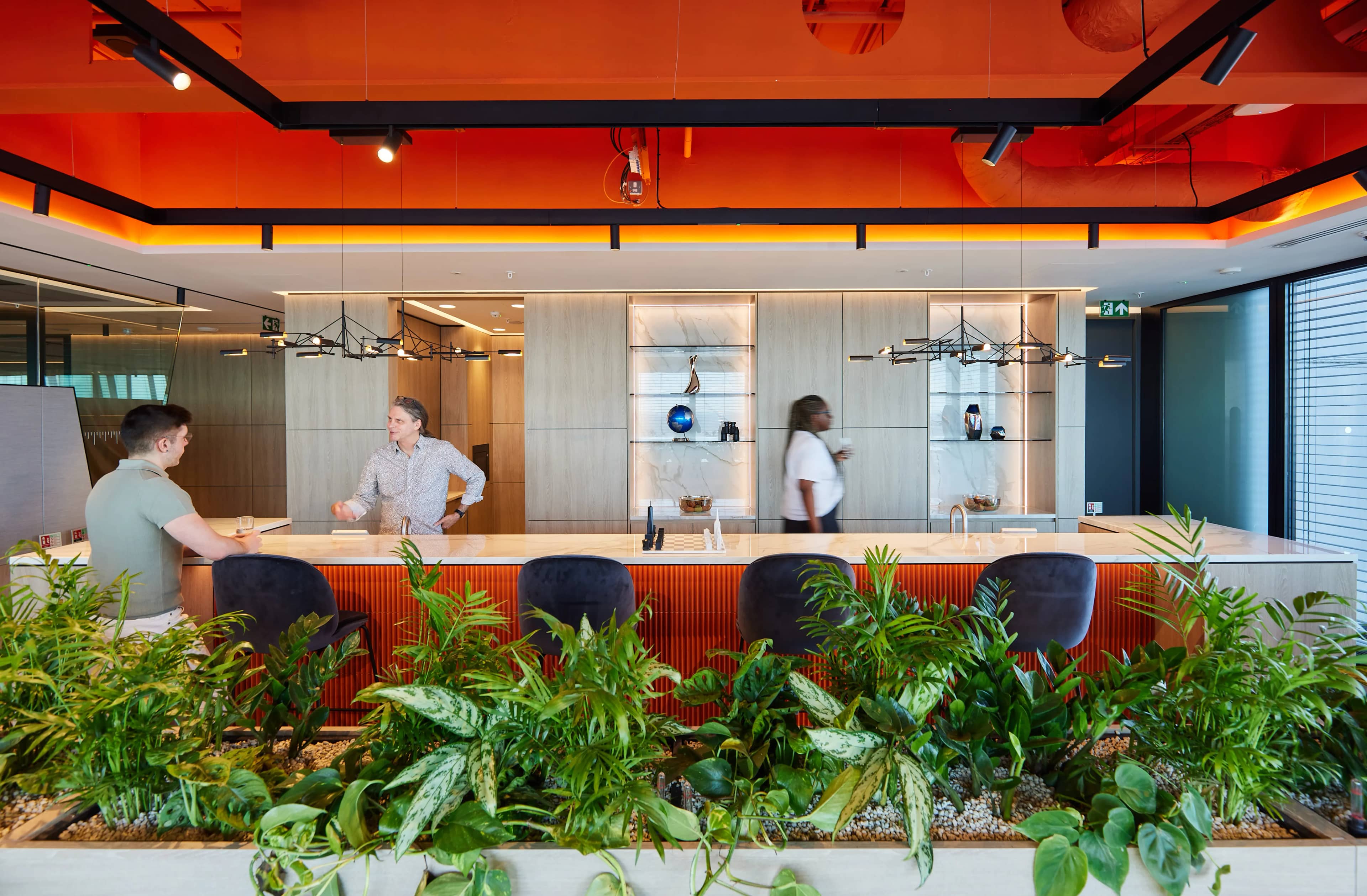 Modern office interior with two people conversing at a counter, surrounded by lush green plants and orange ceiling accents.