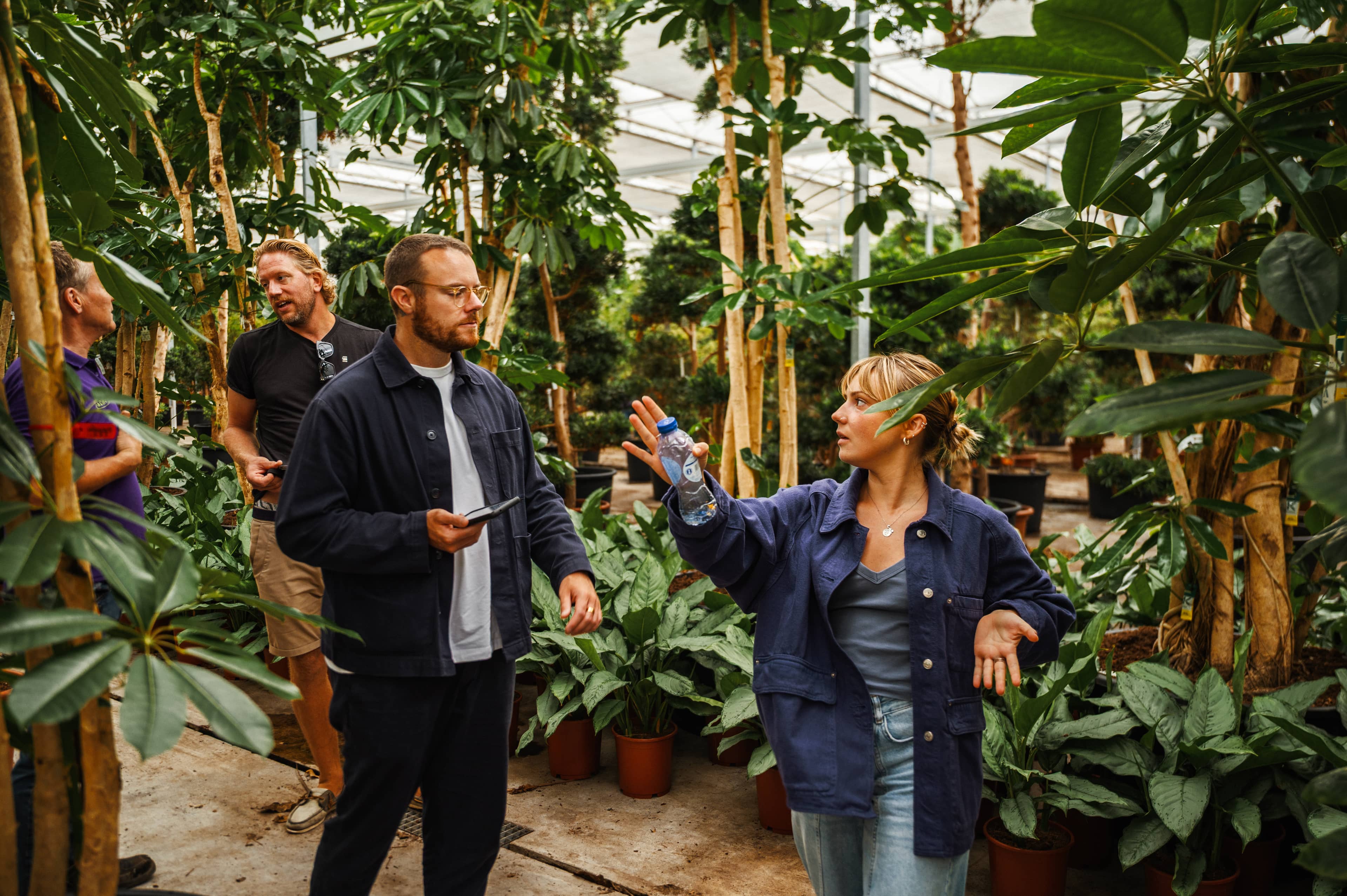 People walking and discussing inside a greenhouse filled with tall plants, ficus trees, and potted greenery.
