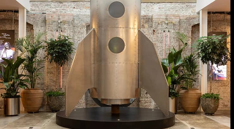 A metallic rocket model with round windows is displayed indoors, surrounded by green plants in large pots, against a brick wall background.