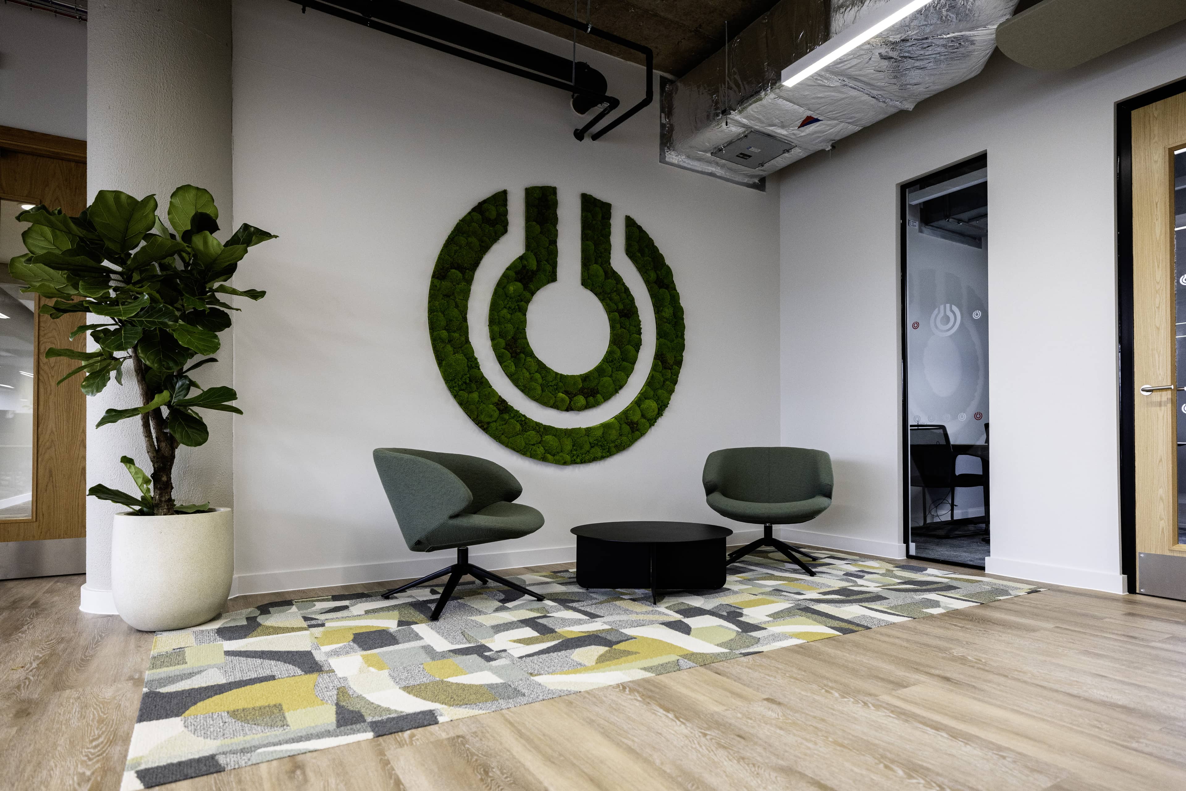 Modern office waiting area with two green chairs, a round black table, a plant, and a moss wall art piece resembling a power symbol.