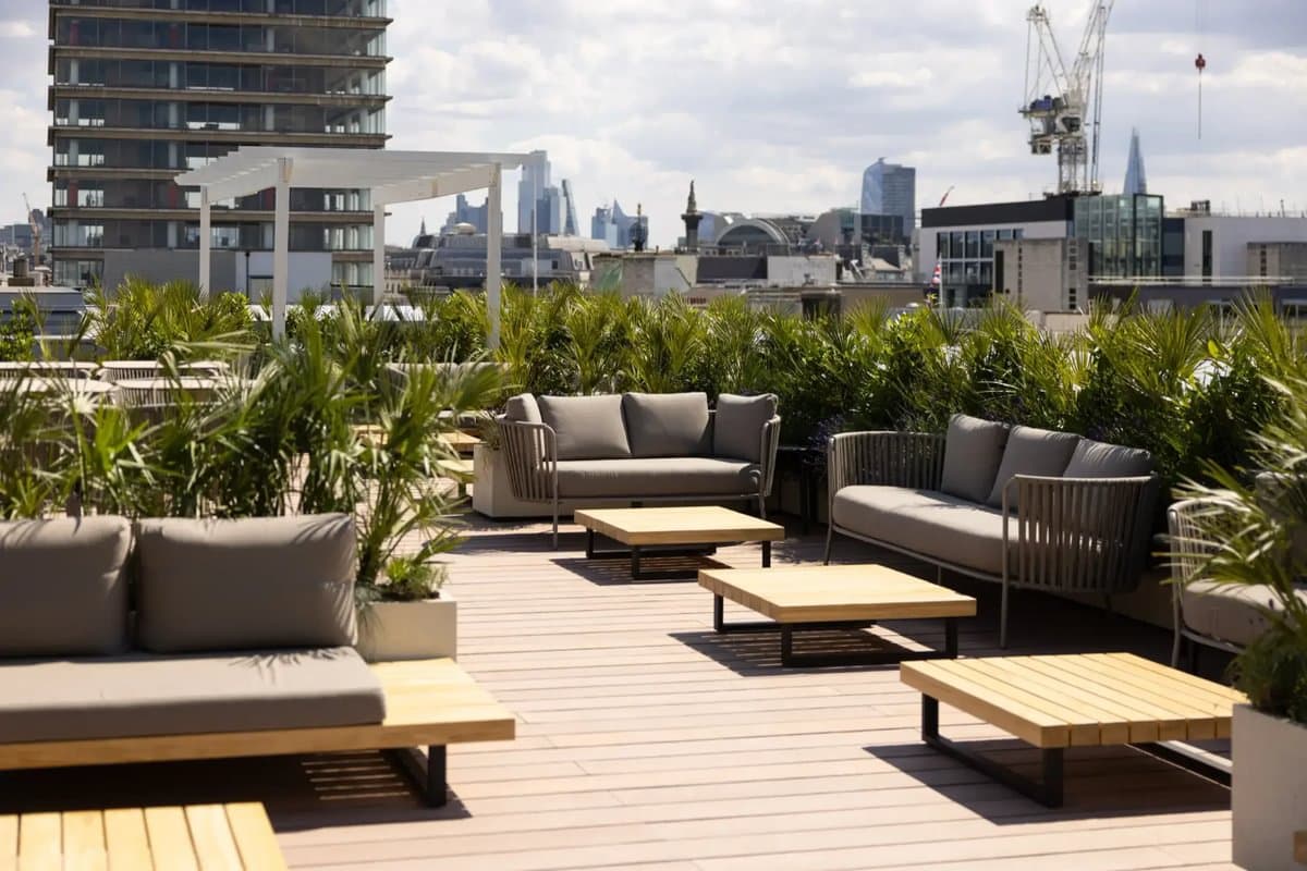 Rooftop patio with modern sofas, coffee tables, and surrounding greenery, overlooking city skyline on a partly cloudy day.